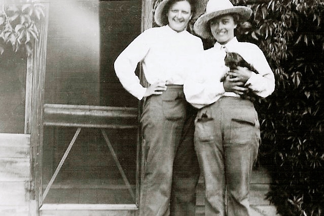 Black and white photo of two women standing outside a screen door. Both are wearing work trousers, white shirts, and large hats. The woman on the right is holding a cat.