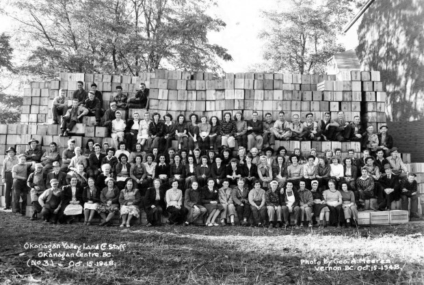 Black and white photo of a large group of men and women outside. Most are sitting on apple boxes stacked in tiers.