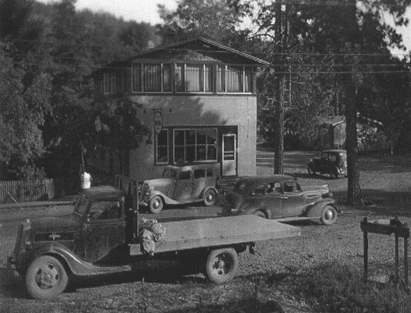 Black and white photo of a building with an older model truck and three older model cars.