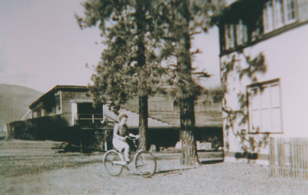 Black and white photo of a young woman riding a bicycle. A wooden building with a loading door is in the background.