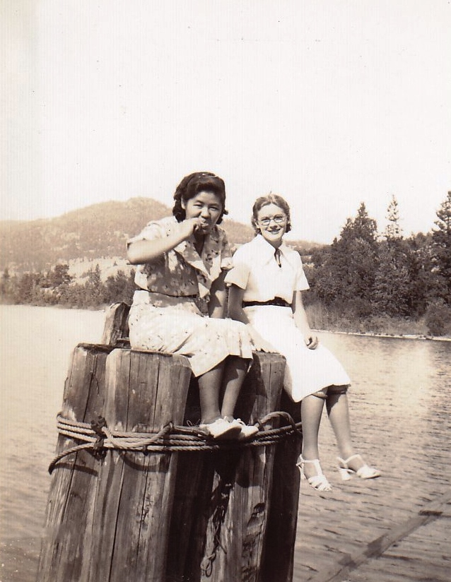 Black and white photo of two young women sitting on pilings over a lake.
