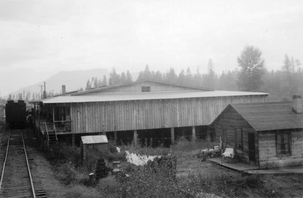 Black and white photo of a wooden house with laundry outside. A large wooden building is next to the house and a railway track runs in front of both buildings.