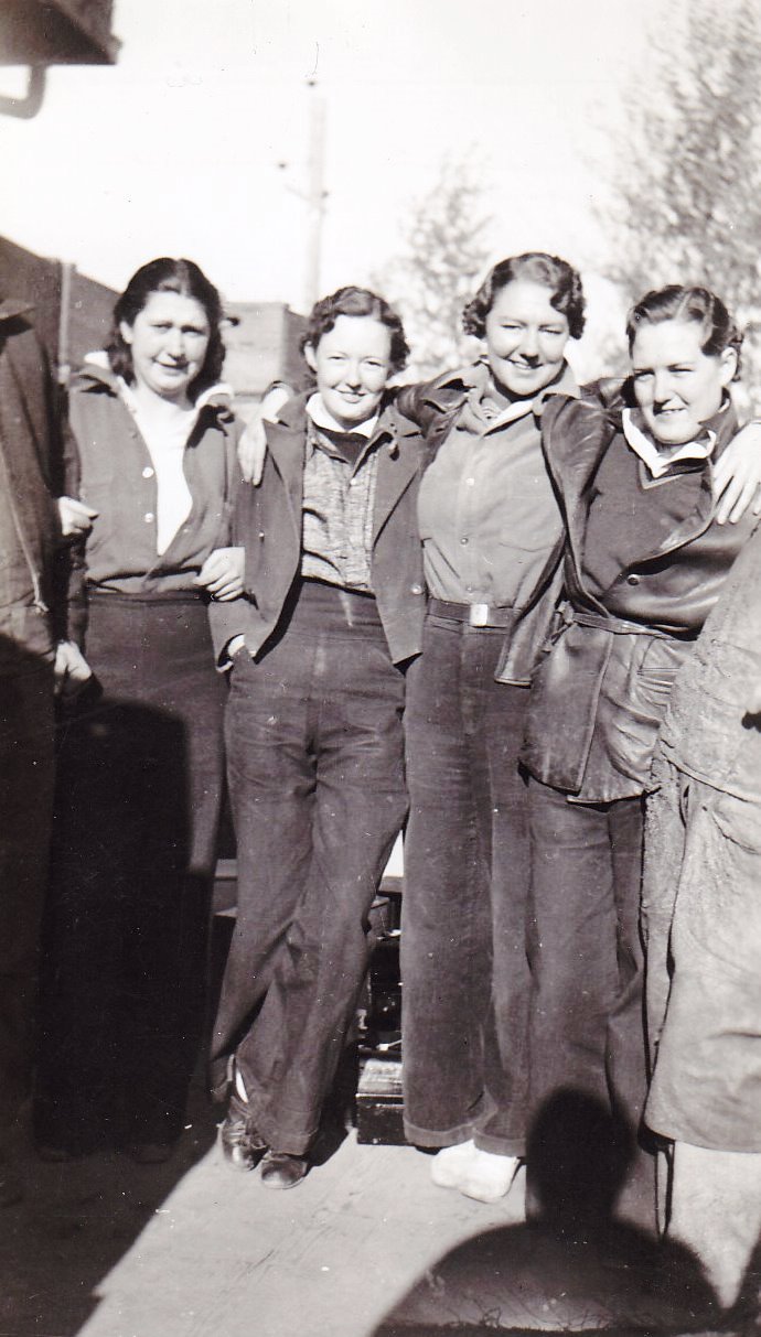 Black and white photo of four women standing outside. They are wearing pants and have their arms across each other's shoulders.