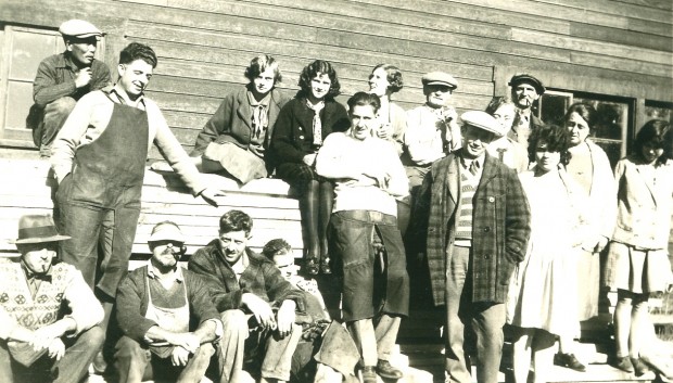 Black and white candid photo of ten men, ages early 20's to mid-40's, and seven young women all in casual clothing outside a wooden building. Some are sitting on stacked lumber; others are standing or leaning back. Two men are smoking pipes.