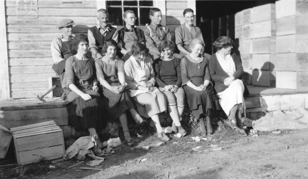 Black and white photo of six women (front) and five men (back) seated in front of an old building. They are wearing 1920's style clothing.