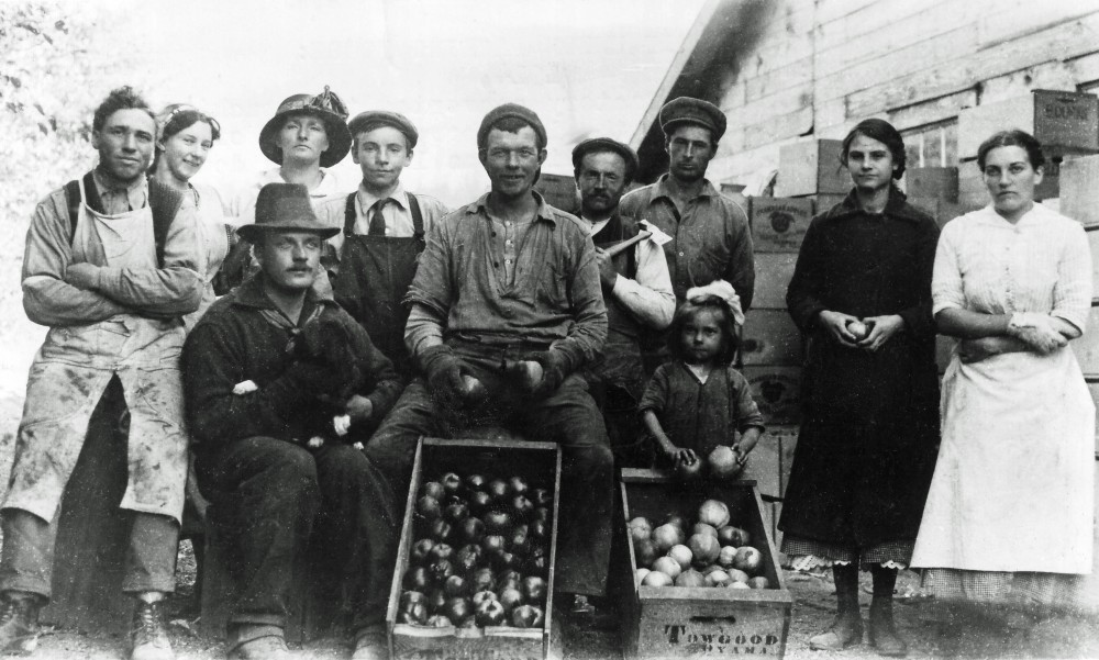 Black and white photo of six men, four women, and one very young girl outside. Behind them is a wooden building. Two wooden boxes of apples are in the foreground; one of the boxes is stamped 