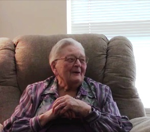 Colour photo of a senior woman with white hair and glasses sitting in a chair, looking to her left and smiling. She is wearing a lavender striped blouse.