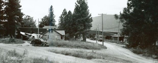 Black and white photo of an old car in front of a wooden house.