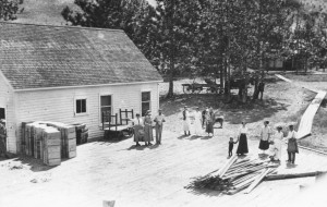 Black and white photo of several men, women, and children standing on a large wooden patio in front of a white wooden building, all wearing early 1900's style clothing. To the left are stacked apple boxes and a cart. In the background are trees, grass, a narrow wooden sidewalk, and a horse and buggy.