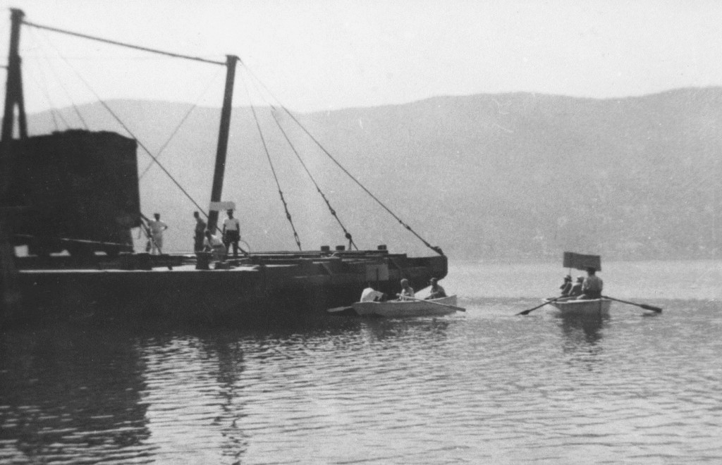 Black and white photo of a barge on which four men are standing. Two small rowboats near the barge each carry three people. Three people are holding up signs.