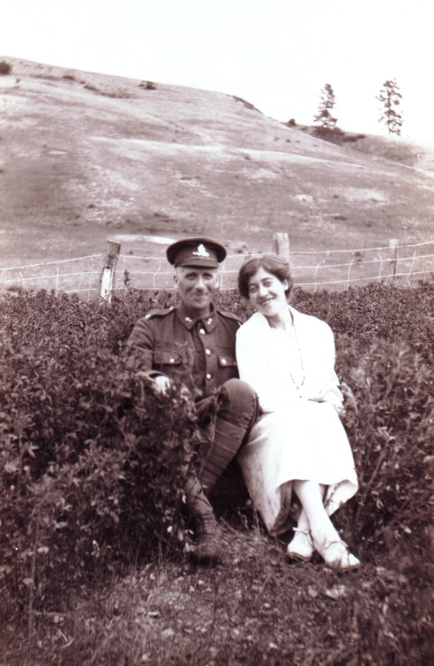 Black and white photo of a man in army uniform and a smiling woman seated outside in a field, with hills in the background.
