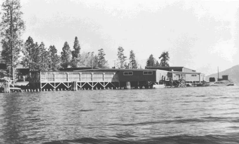 Black and white photo of wooden buildings on the shore of a lake. Sections on pilings extend over the water.