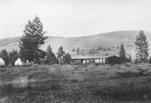 Black and white photo of an old one-story building in a field with hills behind.