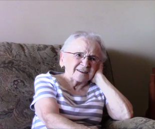 Colour photo of a senior woman with white hair and glasses, sitting in a chair. She is smiling and is resting her head on her left hand.