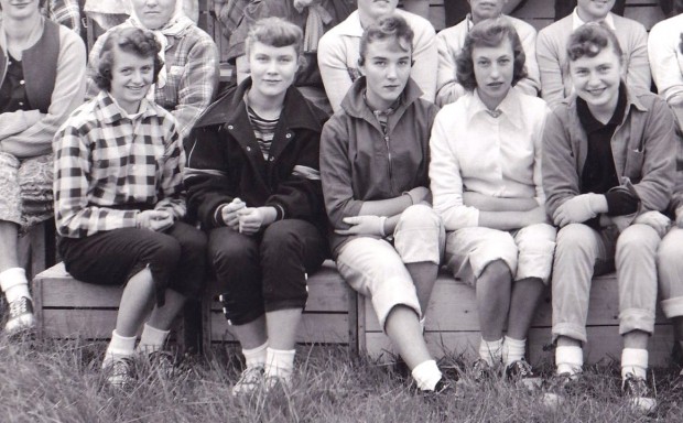 Section of a black and white photo showing five young women seated outside on wooden boxes.