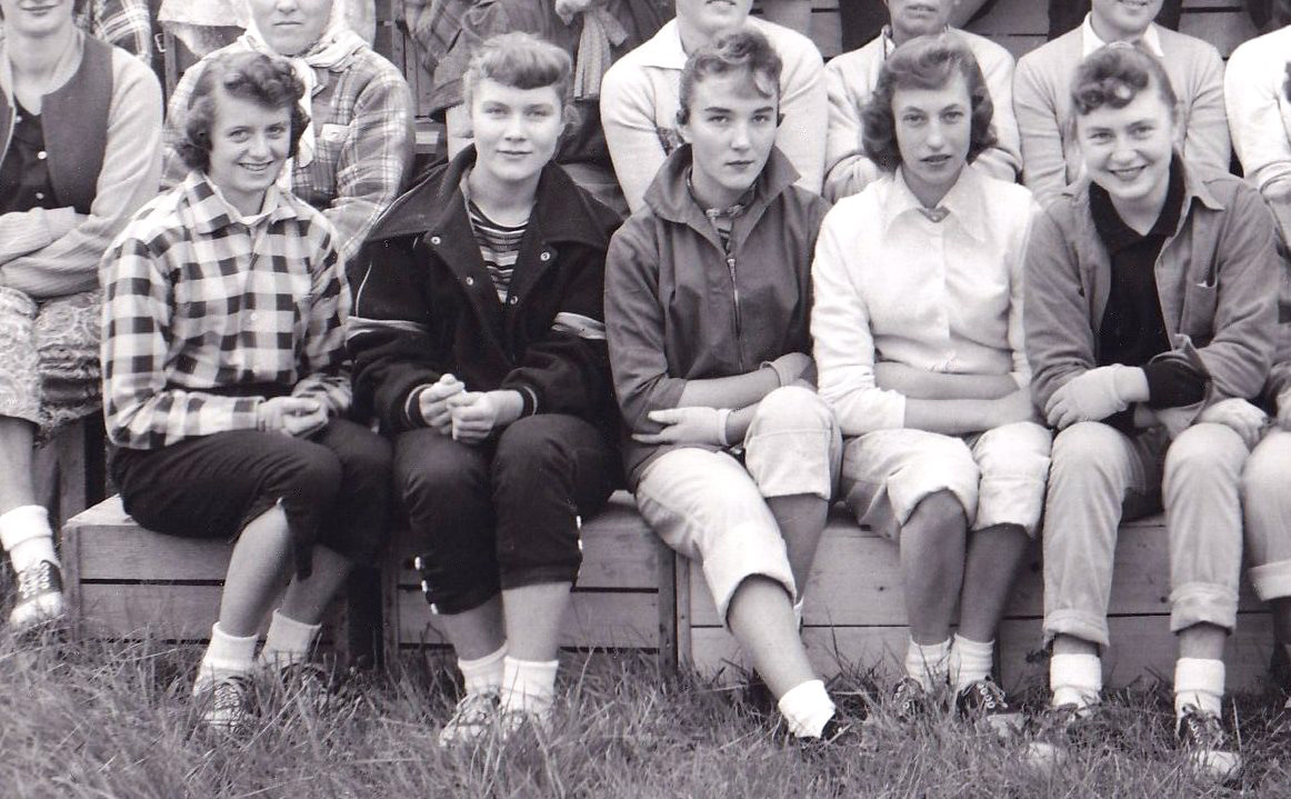 Section of a black and white photo showing five young women seated outside on wooden boxes.