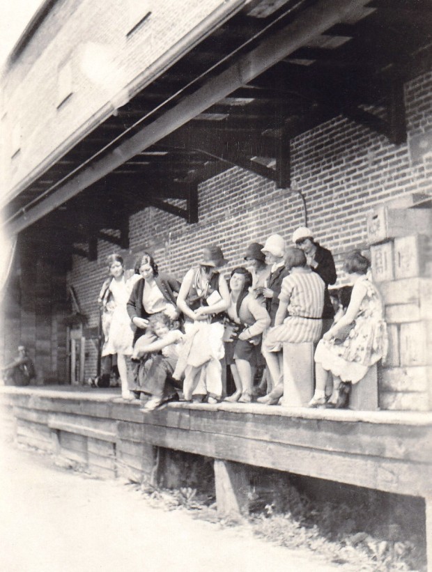 Black and white candid photo of ten women standing or sitting on a deck in front of a brick building.