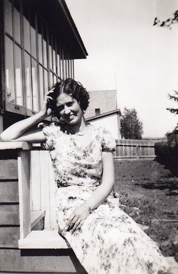 Black and white photo of a young woman in a flowered dress sitting on steps outside a building.