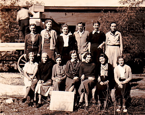 Photo sépia représentant douze femmes et un homme assis ou debout dehors. Derrière eux se dresse un vieil édifice de bois et l’on aperçoit un homme en train de décharger des caisses d’un wagon.