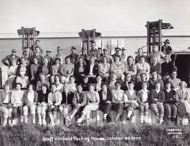 Black and white photo of a large group of men and women seated in tiers outside. Three tall forklifts and a building are behind them.