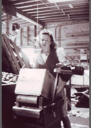 Black and white photo of a young woman standing behind a tilted wooden box. She is inside a building and there is a bin of apples to her right.