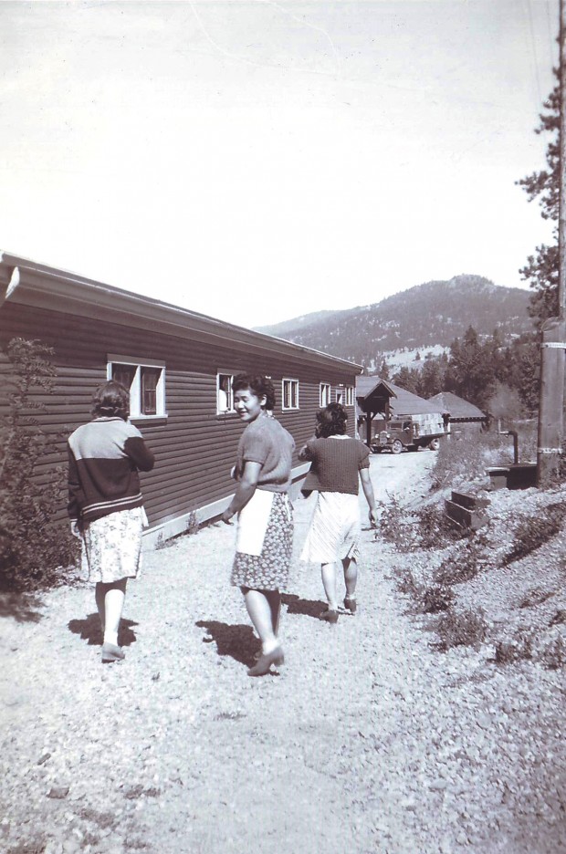Black and white photo of three women walking along a path next to a one-story wooden building. Two women are facing forward, away from the photographer and the third is looking back over her shoulder.