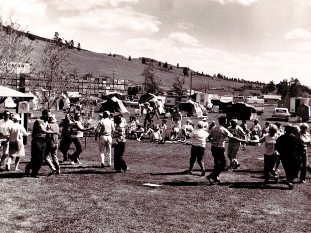 Black and white photo of a large group of men and women dancing in a field, while others, seated, watch. Behind the dancers are old style cars, trailers, tents, and stacks of apple boxes.