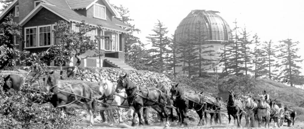 A black and white photograph showing part of the telescope being hauled by horse-drawn wagon with telescope dome and house in background