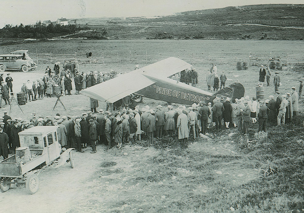 A black and white photograph featuring the 'Pride of Detroit' aircraft on display at airstrip, surrounded by a large crowd. Some automobiles present, as well.