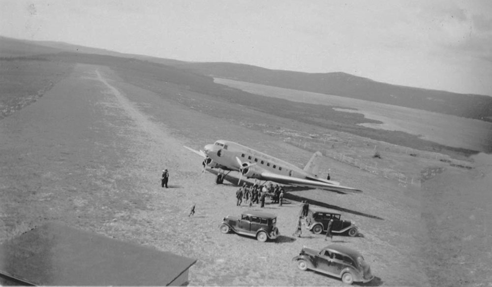 Black and white photo of a Douglas DC-2 plane at the airstrip with some onlookers and three automobiles.