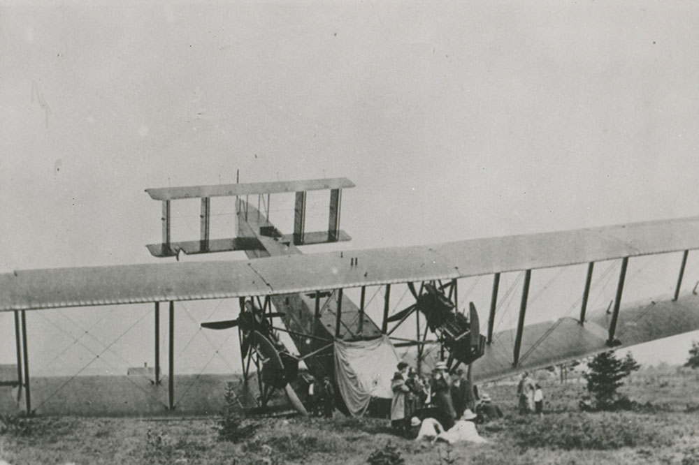 A black and white photograph of the Handley Page Atlantic after crashing in a barren field, nose down with tail in the air. Apparent damage to nose, and right wing. A group of women, finely dressed, are standing in front of the downed aircraft.