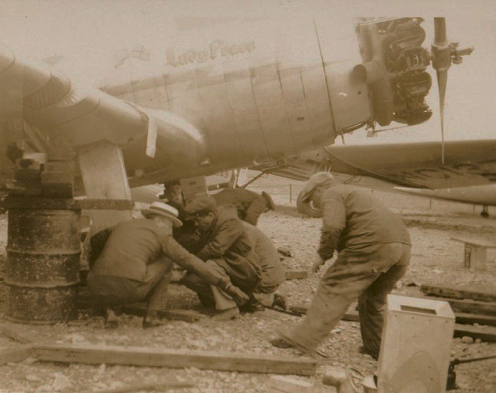Quatre hommes accroupis sous l’avion Lady Peace en train de faire des réparations.