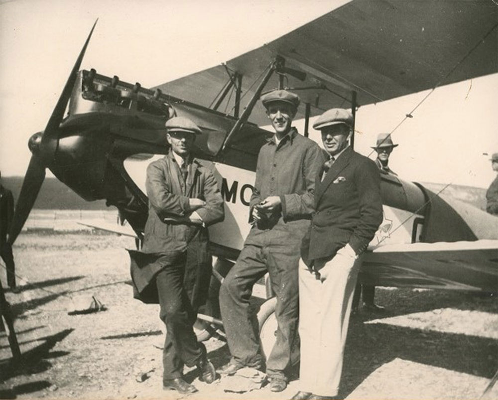A black and white photo of three men stood in front of Gypsy Moth aircraft, with others in background.