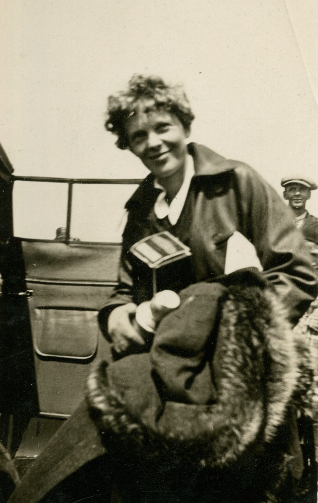 A black and white photograph of Amelia Earhart, in her flight suit, standing next to the open door of a vehicle. She is holding a thermos, a box and a fur lined coat.