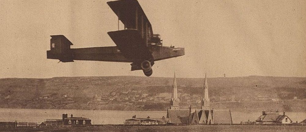 A black and white newspaper photograph of the Handley Page Atlantic flying approximately 20 feet over the ground during a test flight, church steeples in background, harbour, and southside Harbour Grace.