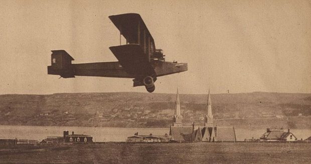 A black and white newspaper photograph of the Handley Page Atlantic flying approximately 20 feet over the ground during a test flight, church steeples in background, harbour, and southside Harbour Grace.