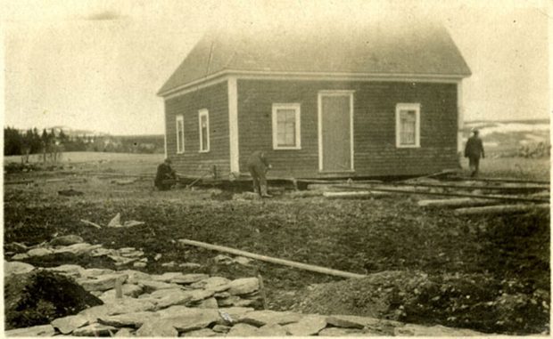 A black and white photo of a house being moved by three men who are placing large logs under the house. Many logs scattered on the ground and flat rocks.