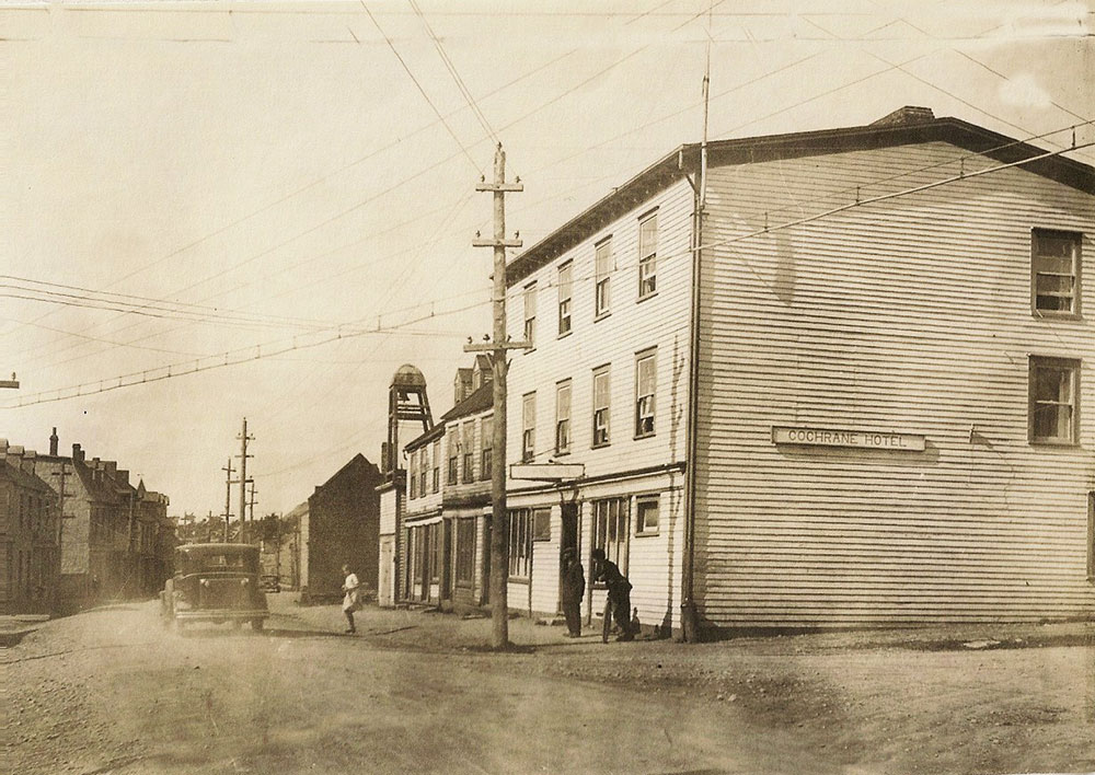 A black and white photo from Water street in Harbour Grace, 1930's, shows a three story building, Cochrane Hotel. A couple of individuals stood in front of the hotel while a vehicle drives down the road.