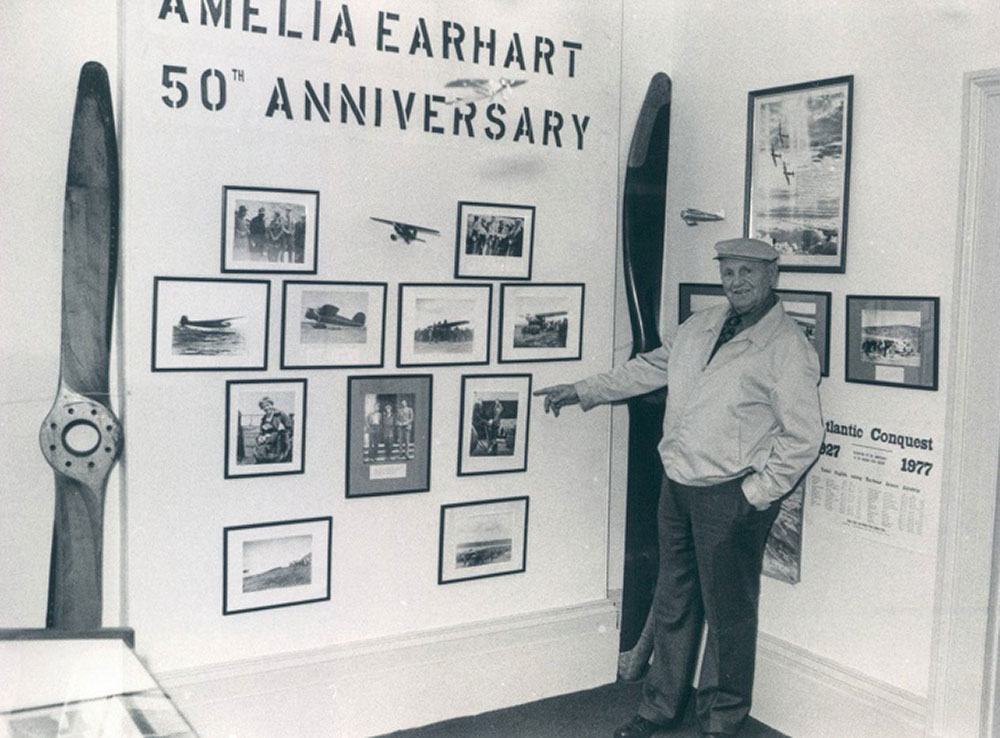 A black and white photograph of Bill Parsons pointing to pictures of Amelia Earhart on display at the Conception Bay Museum. There are two plane propellers on display in room.