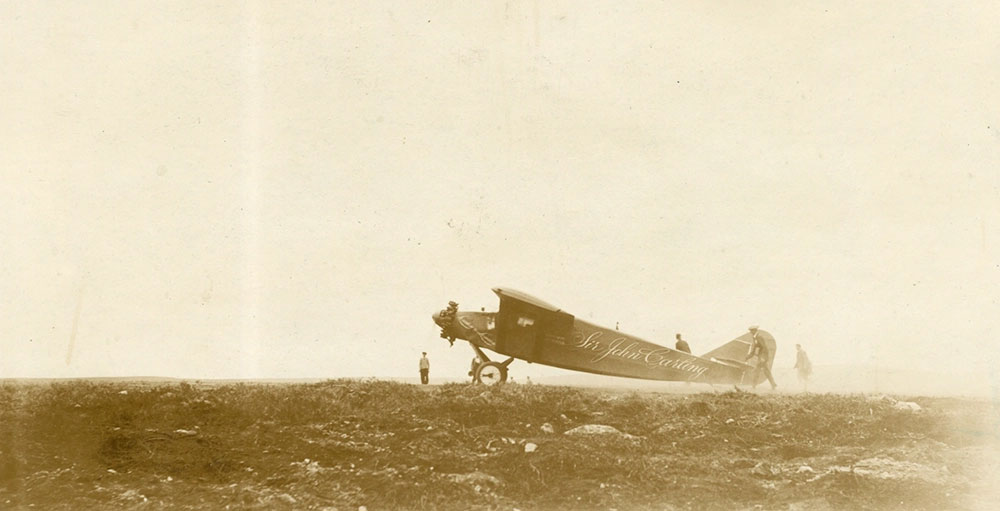 Black and white photo, side view of the Sir John Carling, secure at airstrip, from a distance surrounded by thick fog. Four individuals standing around the aircraft.