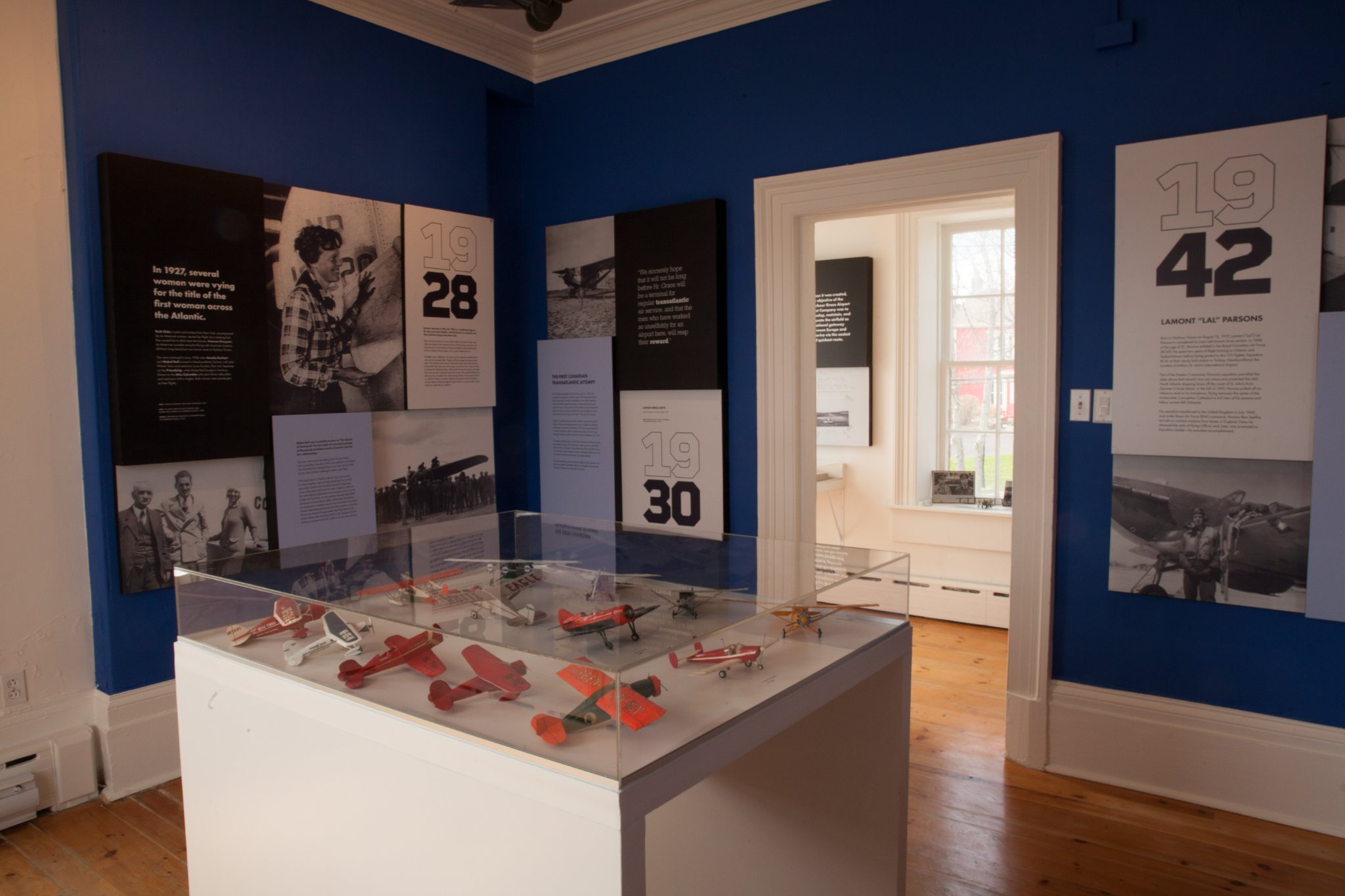 A coloured photograph of an exhibit space with storyboards posted on the walls and a glass case displays 12 small model planes.