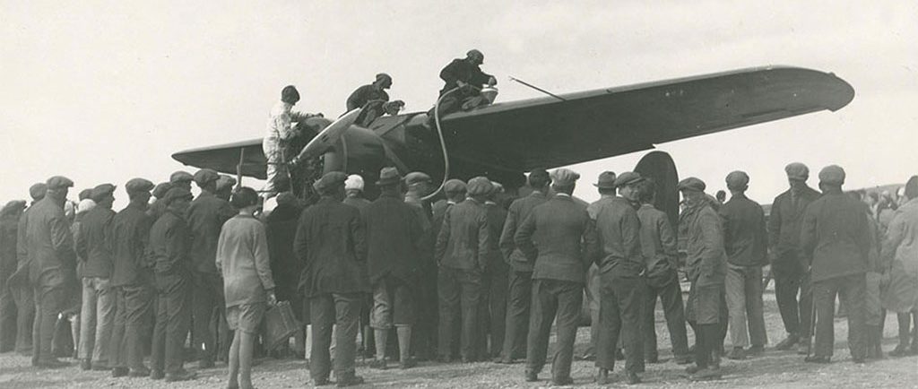 A black and white photo of three individuals on top of the wings of a plane, refueling. A large crowd stood around the aircraft, backs to camera.