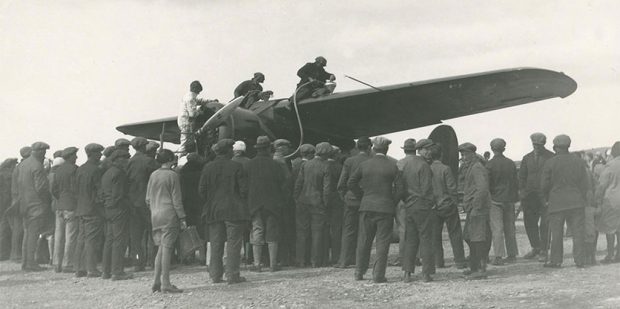 A black and white photo of three individuals on top of the wings of a plane, refueling. A large crowd stood around the aircraft, backs to camera.