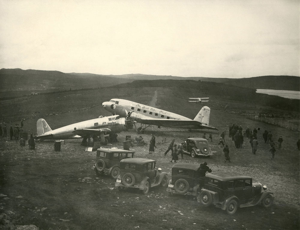 A black and white photograph showing three aircrafts parked at airstrip. Five automobiles parked there with a small crowd gathered.