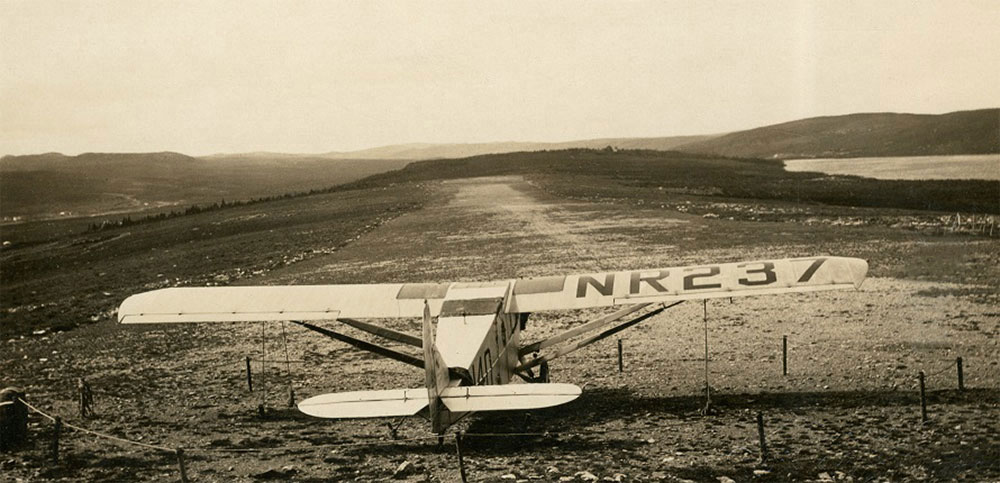 A black and white photograph of the Columbia aircraft secured at foot of clay airstrip. Marker number NR237 is visible across right wing of plane.