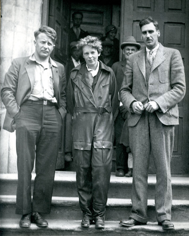 A black and white photograph of Amelia Earhart, in her flight suit, pictured on outside steps of Harbour Grace Post Office, a gentleman on each side, with other people in background.