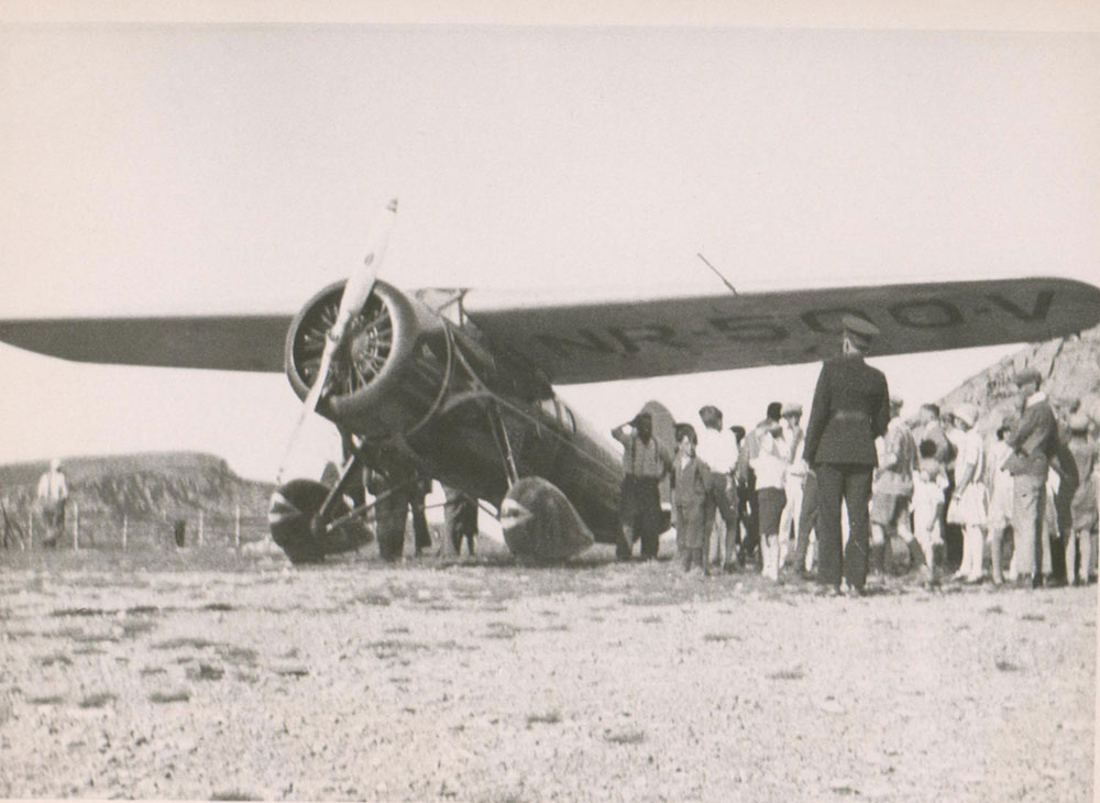 A black and white photograph of the City of New York plane secured at airstrip surrounded by a crowd, including a man in uniform, possibly a policeman. Marker number NR-500-V can be seen under the right wing.