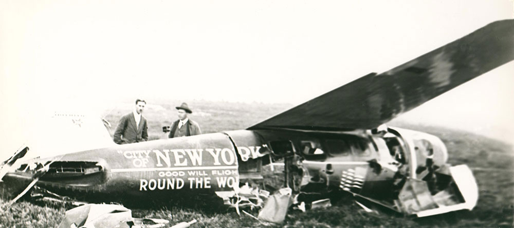 A black and white photograph of two men stood behind the City of New York plane after crashing at Harbour Grace airstrip. Aircraft is extensively damaged on right side in photo.