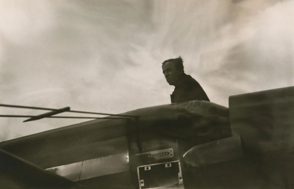 A black and white photo of Pilot James Mollison on top of his aircraft, the Dorothy, at airstrip.