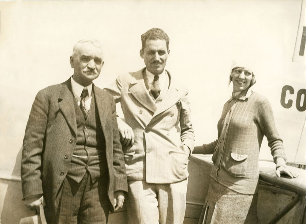 A black and white photograph of three individuals stood in front of the Columbia aircraft. Mabel Boll far right, leaning on the plane's wing. Two of the men are wearing suits. Ms. Boll is wearing a white flight hat with a sweater and skirt.